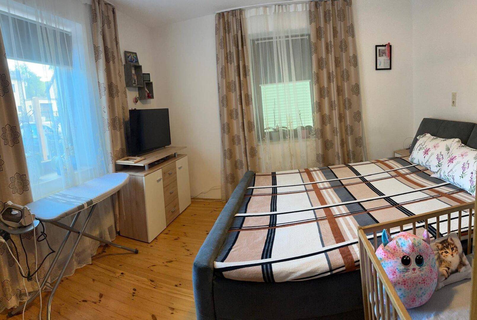 Schlafzimmer mit Doppelbett, Babybett und Holzboden, hell durch zwei Fenster.