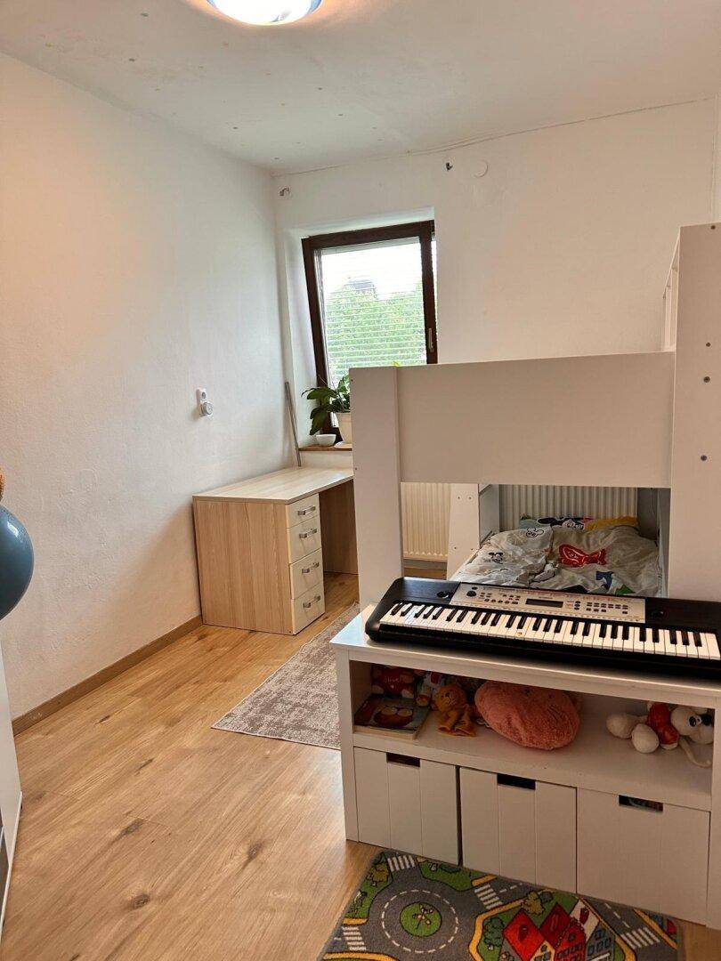 Kinderzimmer mit Hochbett, Schreibtisch und Holzboden, hell durch ein Fenster.