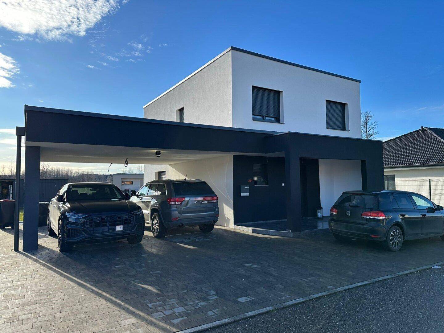 Modernes Einfamilienhaus mit weißer Fassade, schwarzem Carport und gepflasterter Einfahrt.
