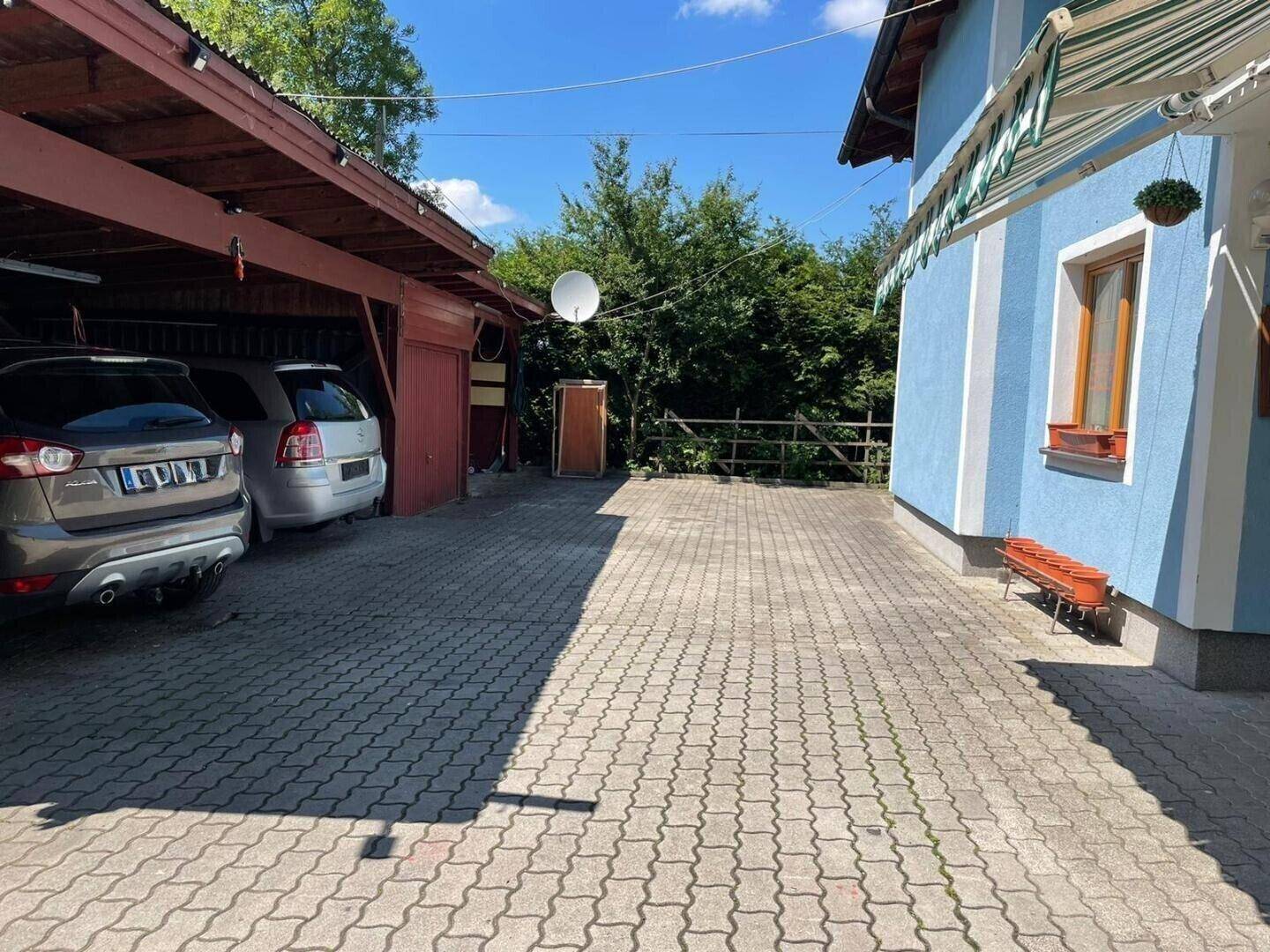 Außenansicht eines Hauses mit blau-weißer Fassade, angrenzendem Carport und gepflasterter Einfahrt unter blauem Himmel.