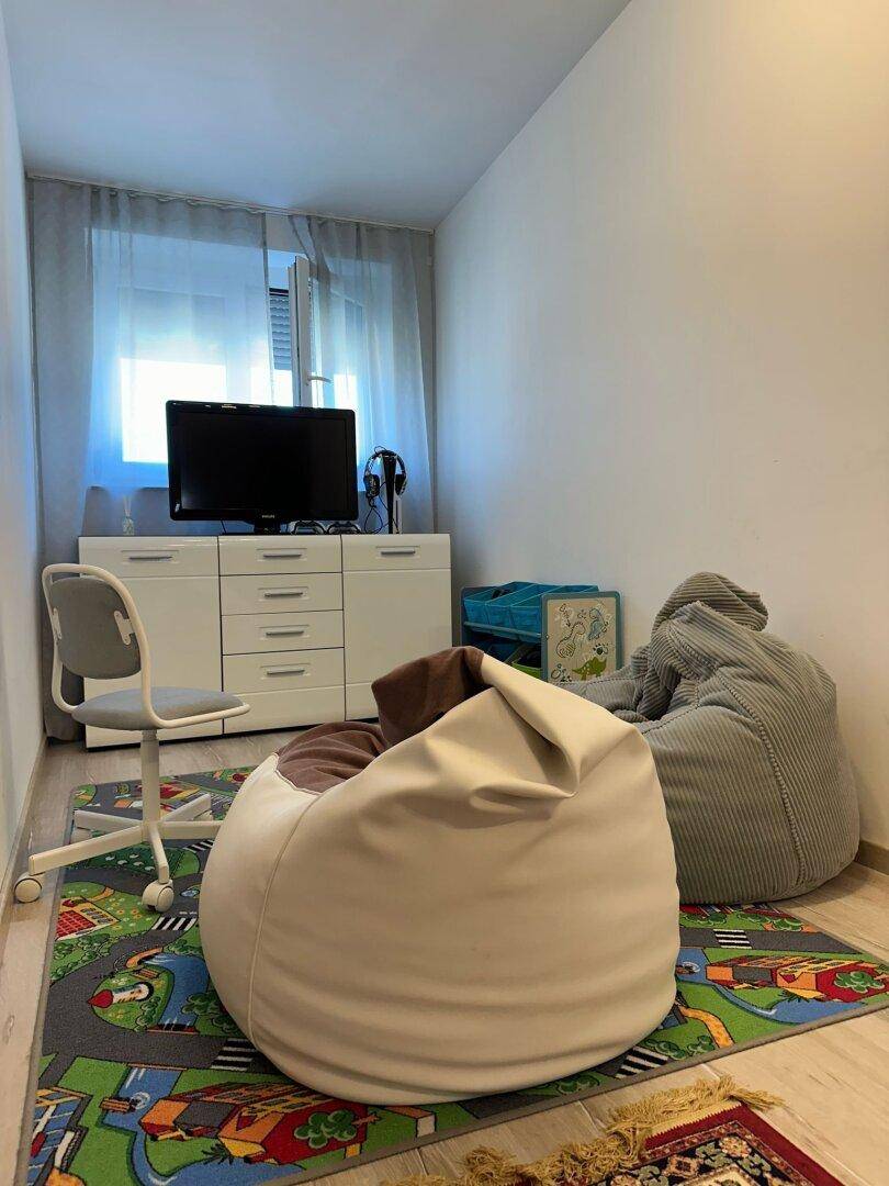 Kleines Zimmer mit Fernseher, Schreibtisch und zwei Sitzsäcken auf einem bunten Teppich.