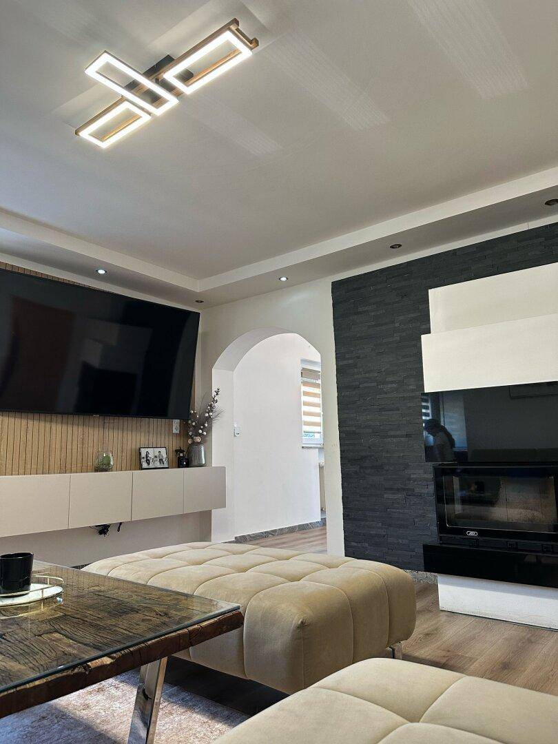 Modernes Wohnzimmer mit Kamin, großem Fernseher und beigem Polsterhocker unter einer Deckenleuchte.
