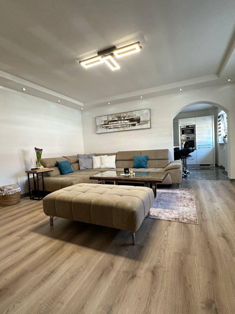 Geräumiges Wohnzimmer mit beigem Ecksofa, Holzboden und Blick in die angrenzende Küche.