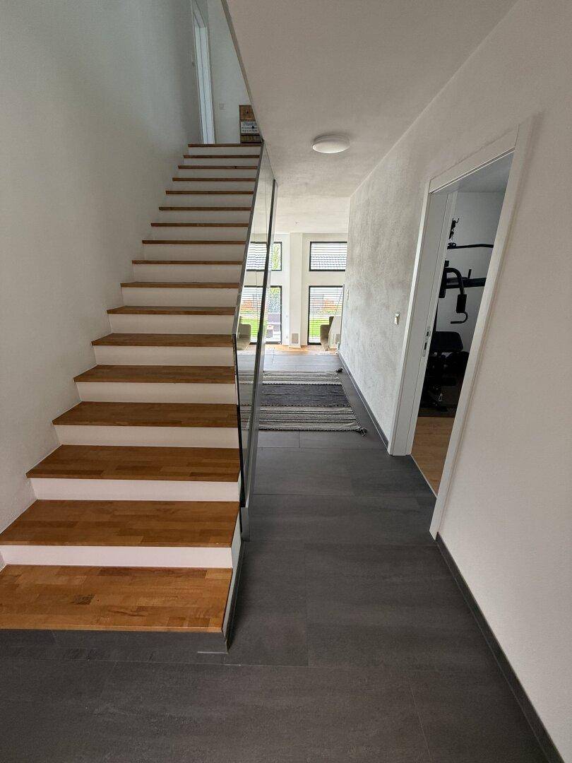 Heller Flur mit Holztreppe, Glasgeländer und dunklem Fliesenboden, der zu weiteren Räumen führt.