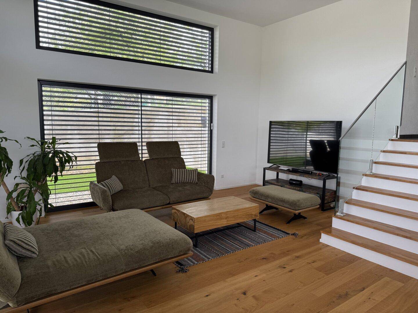 Gemütliches Wohnzimmer mit Holzboden, bequemen Sofas und großen Fenstern mit Jalousien.