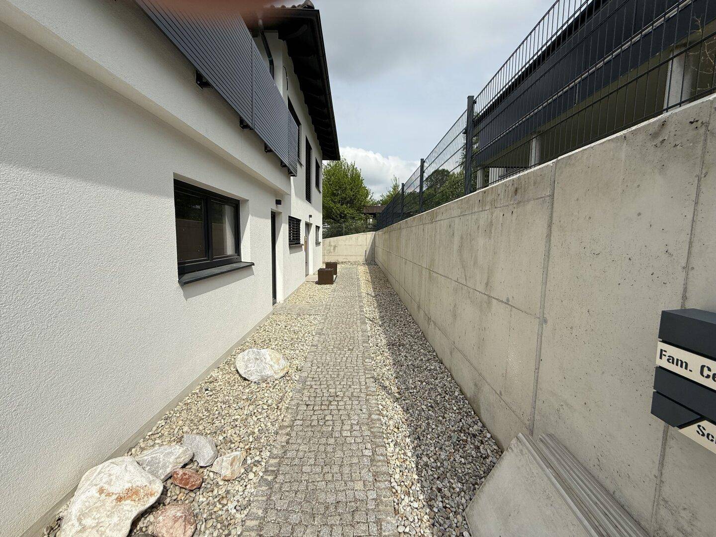 Schmaler gepflasterter Weg entlang der Hauswand mit Kiesbeet und Betonmauer.