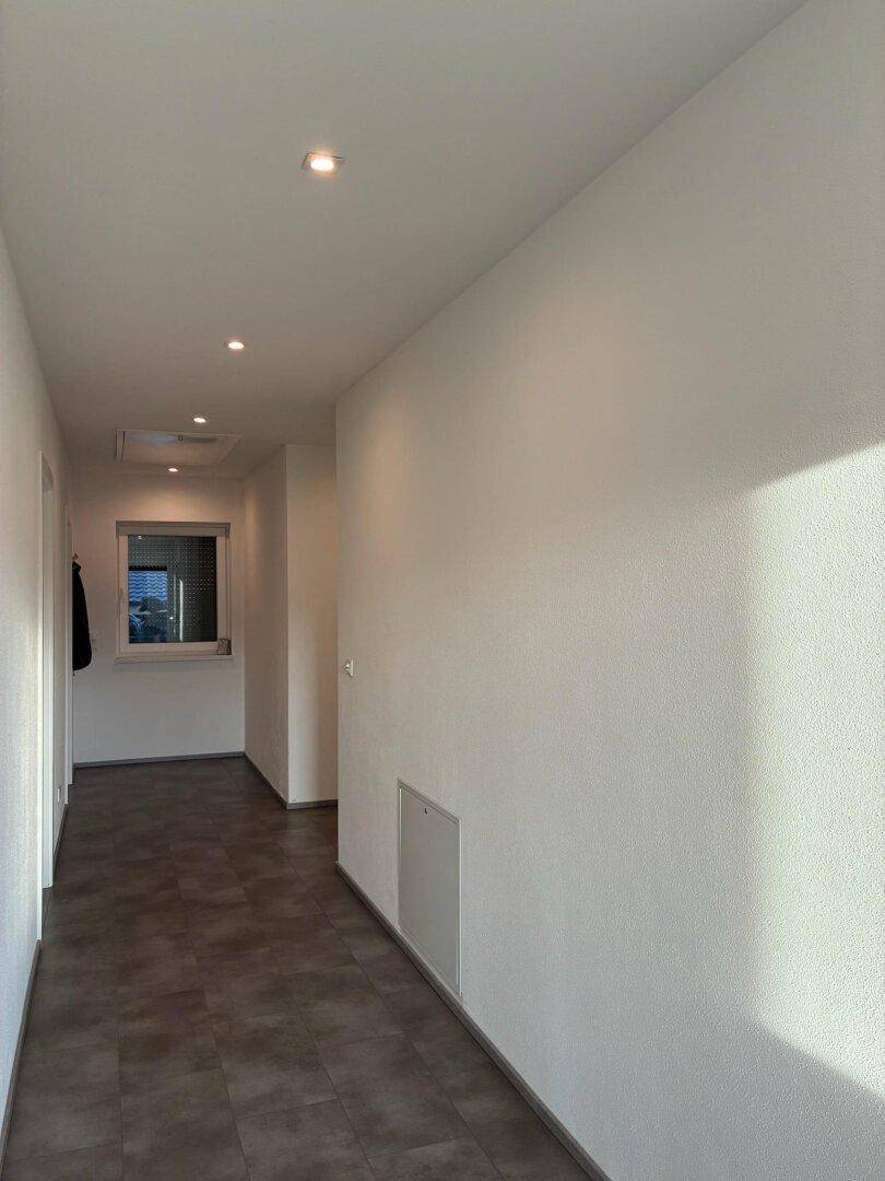 Langer, heller Flur mit Fliesenboden und einem Fenster am Ende.