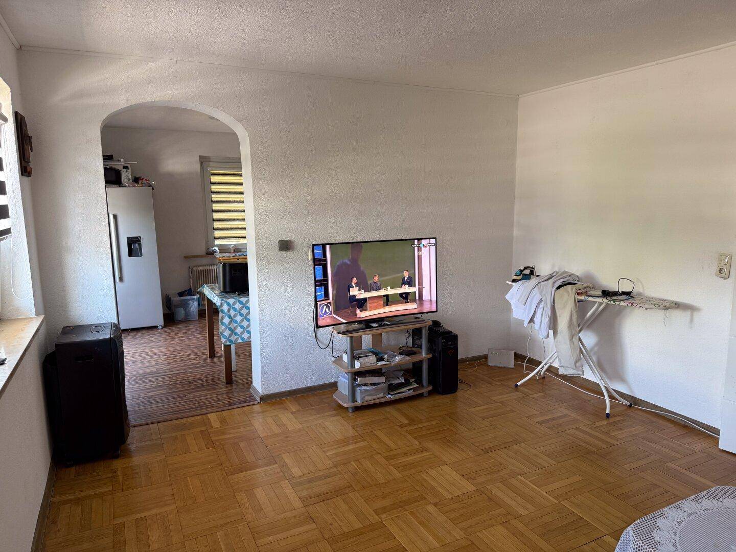 Heller Wohnbereich mit Fernseher und offenem Übergang zum Essbereich durch einen Rundbogen.