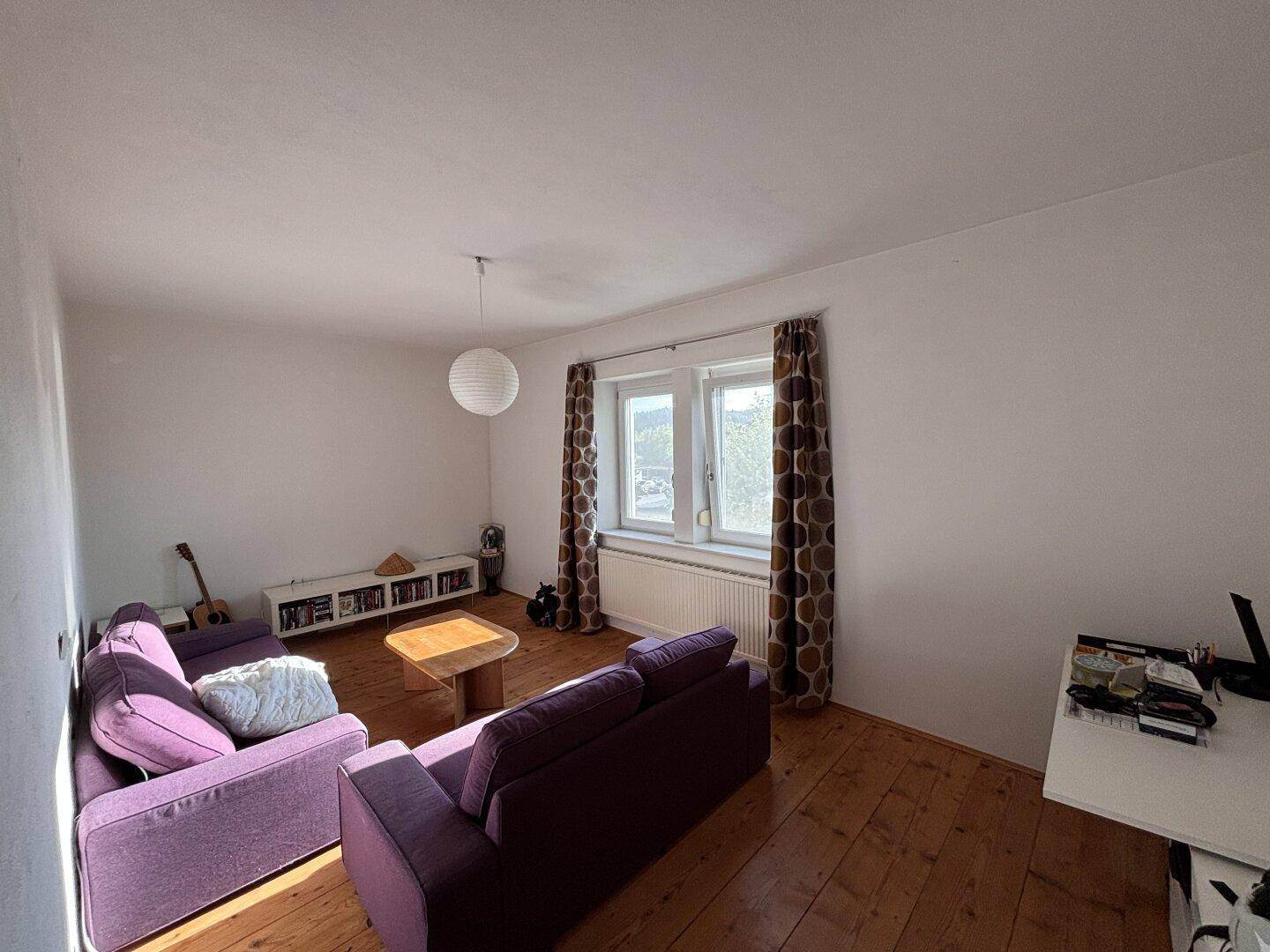 Helles Wohnzimmer mit Holzdielenboden, bequemen lila Sofas und Blick ins Grüne.