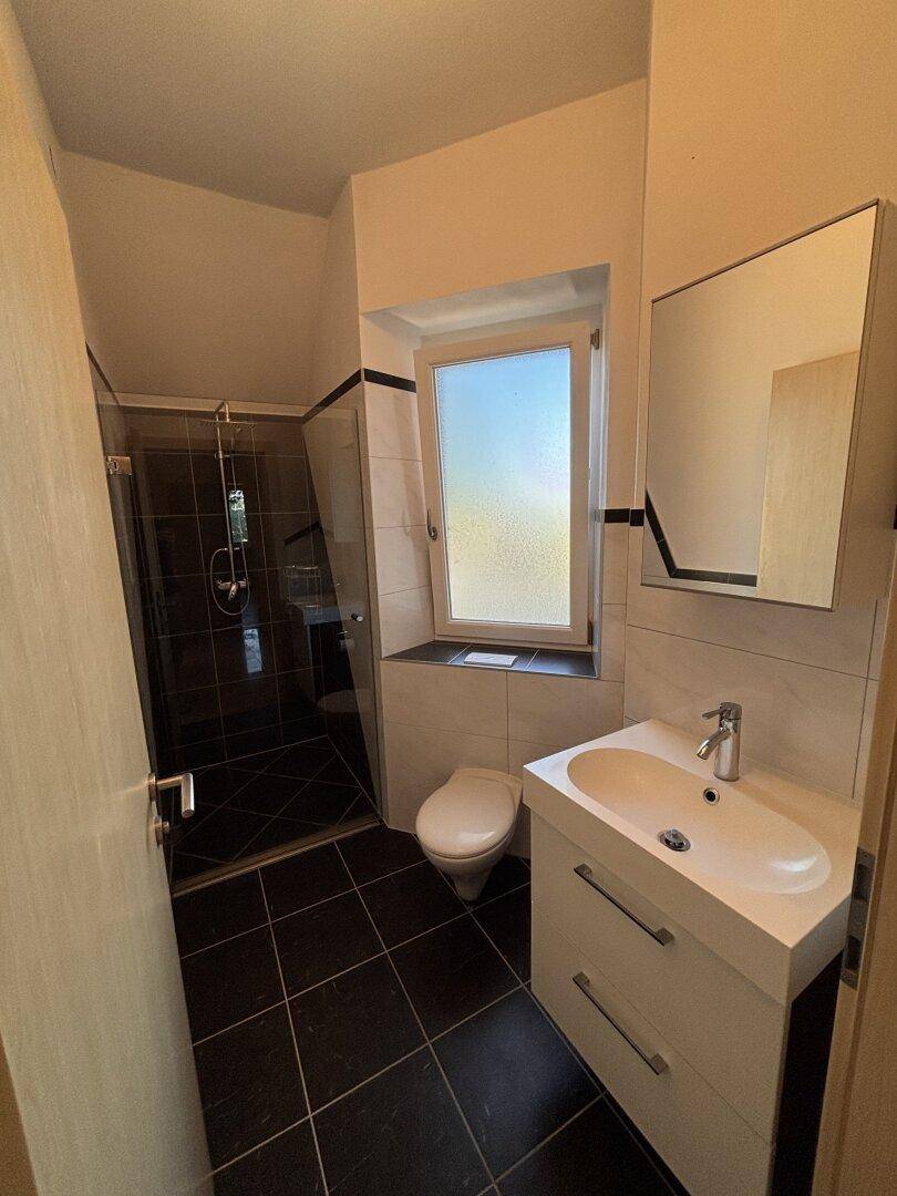 Modernes Badezimmer mit dunklen Fliesen, ebenerdiger Dusche und Fenster für Tageslicht.