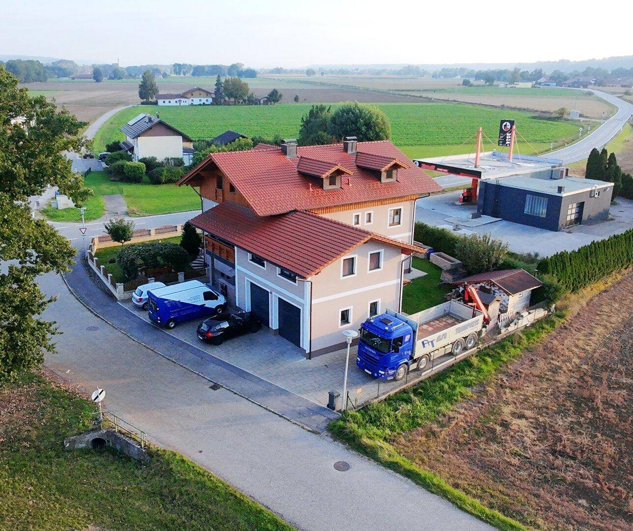 Weitläufige Luftaufnahme eines Hauses mit rotem Dach, umgeben von Feldern und einer Tankstelle.