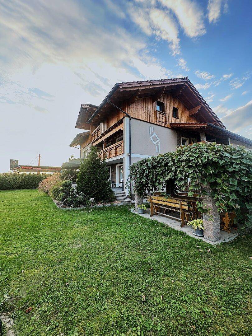 Seitlicher Blick auf das Haus mit Holzfassade, gepflegtem Rasen und einer überdachten Sitzbank im Garten.