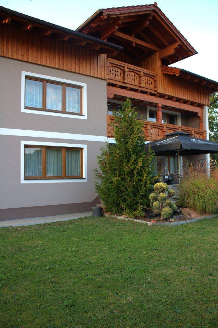 Seitenansicht des Hauses mit Holzfassade, gepflegtem Rasen und einer kleinen Terrasse mit Sonnenschirm.