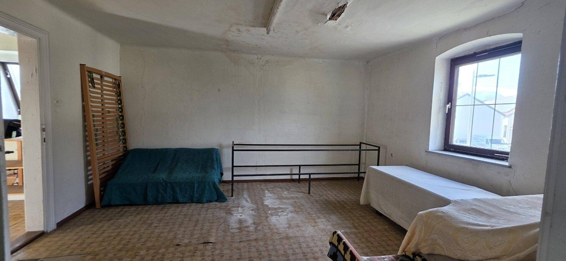 Helles Zimmer mit Fenster, einfacher Einrichtung und deutlichen Renovierungsbedarf.