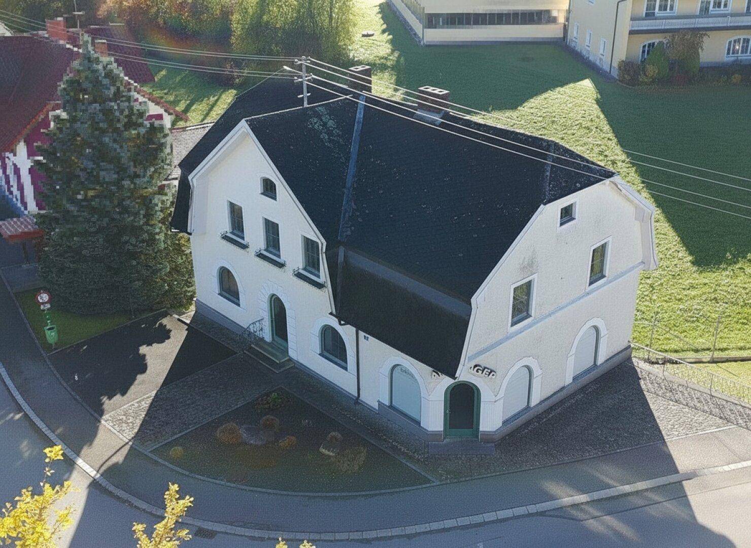 Luftaufnahme des Gebäudes mit dunklem Dach und umliegender Grünfläche bei Sonnenschein.