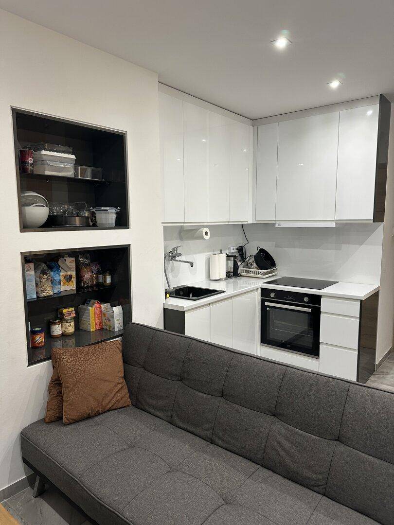 Kompakte, moderne Küchenzeile mit weißen Hochglanzfronten und integriertem Backofen neben einem Sofa.