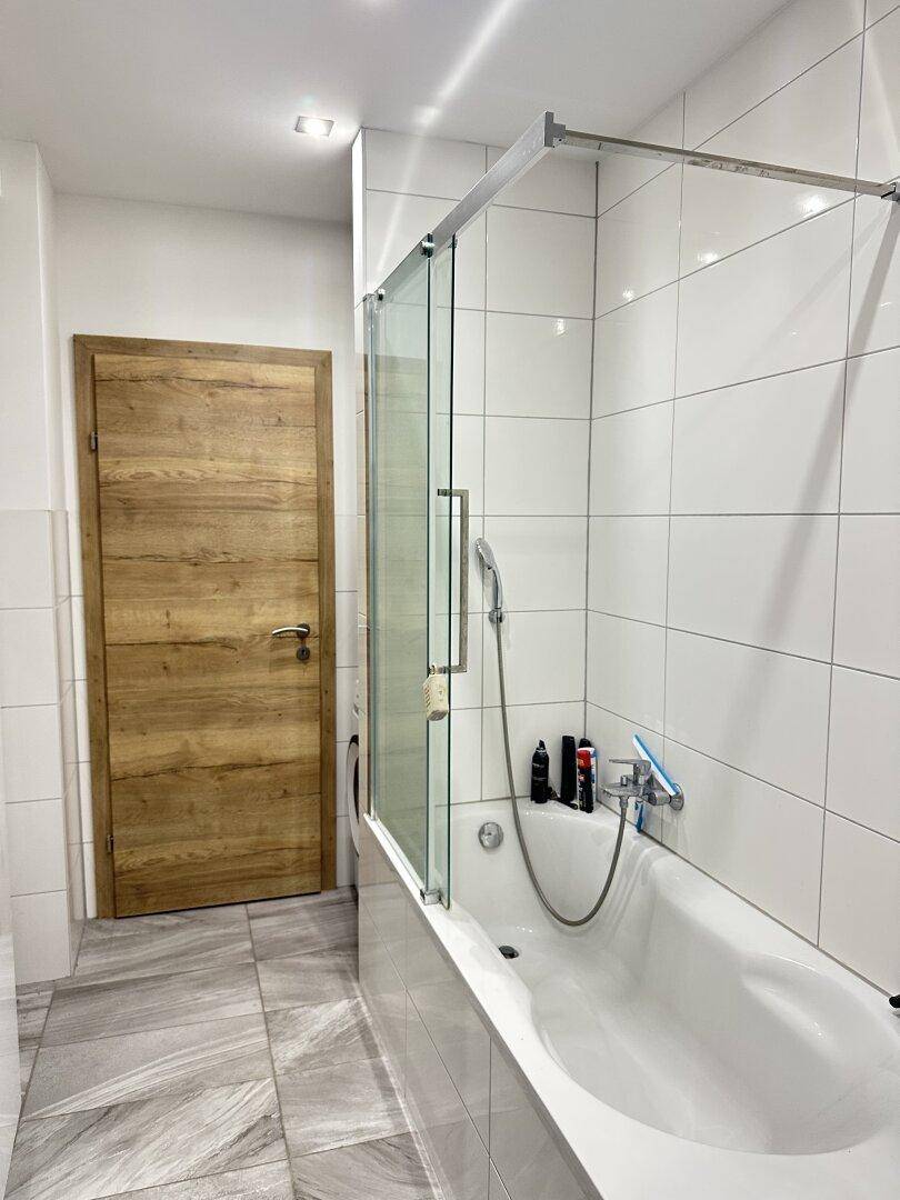 Modernes Badezimmer mit Badewanne und Duschabtrennung, weißen Fliesen und Holztür.