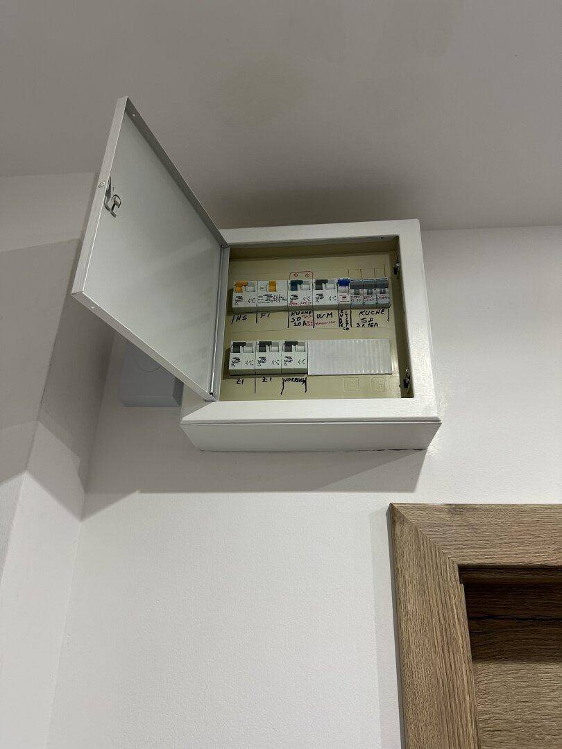 Offener Sicherungskasten an einer weißen Wand, neben einer Holztür.