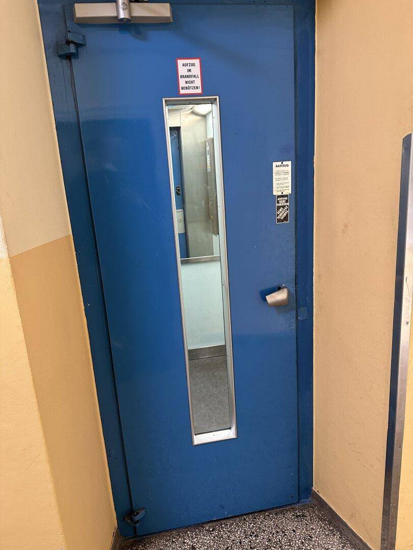 Blaue Aufzugstür in einem Flur mit einem schmalen Fenster und Terrazzoboden.