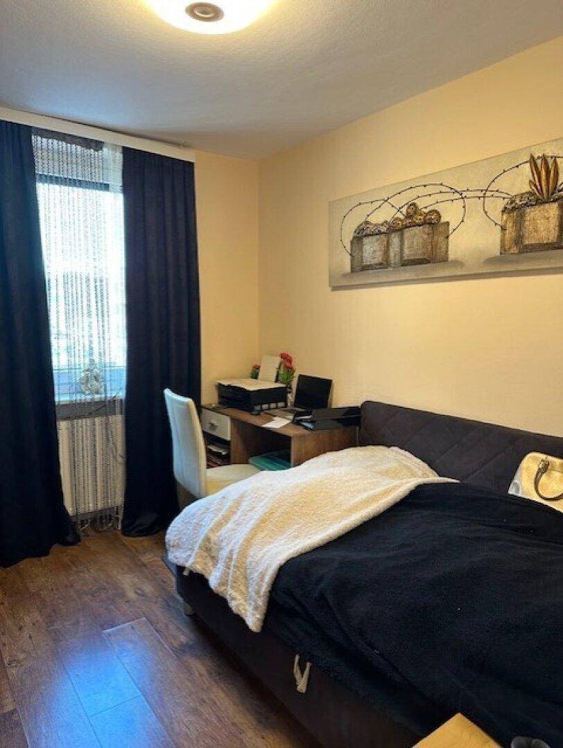 Schlafzimmer mit Bett, Schreibtisch und dunklen Vorhängen am Fenster.