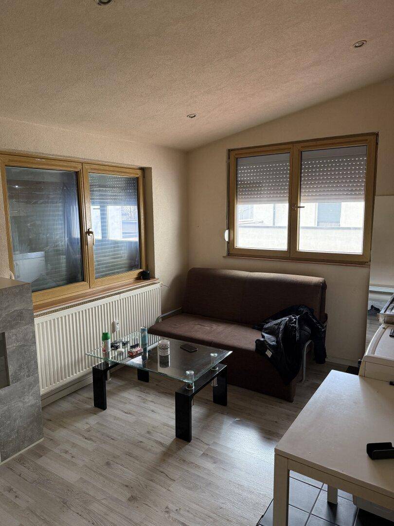 Wohnbereich mit hellem Laminatboden, Sofa und zwei Fenstern mit Jalousien.