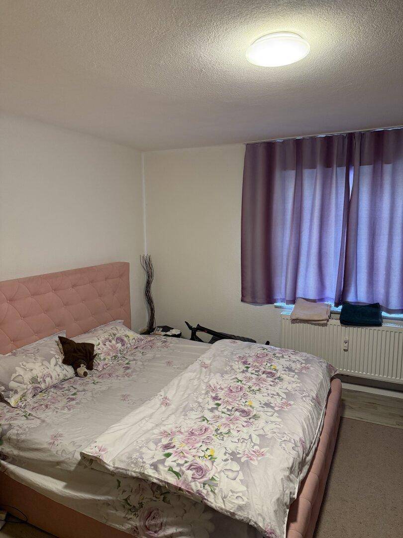 Schlafzimmer mit rosa gepolstertem Bett, hellem Boden und lila Vorhängen am Fenster.