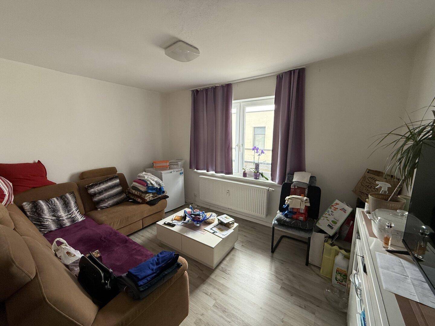 Helles Wohnzimmer mit beigem Sofa, Couchtisch und einem Fenster mit lila Vorhängen.