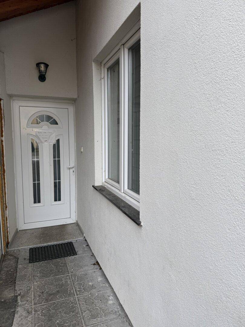 Eingangsbereich des Gebäudes mit weißer Tür und einem Fenster, geschützt durch ein Vordach.