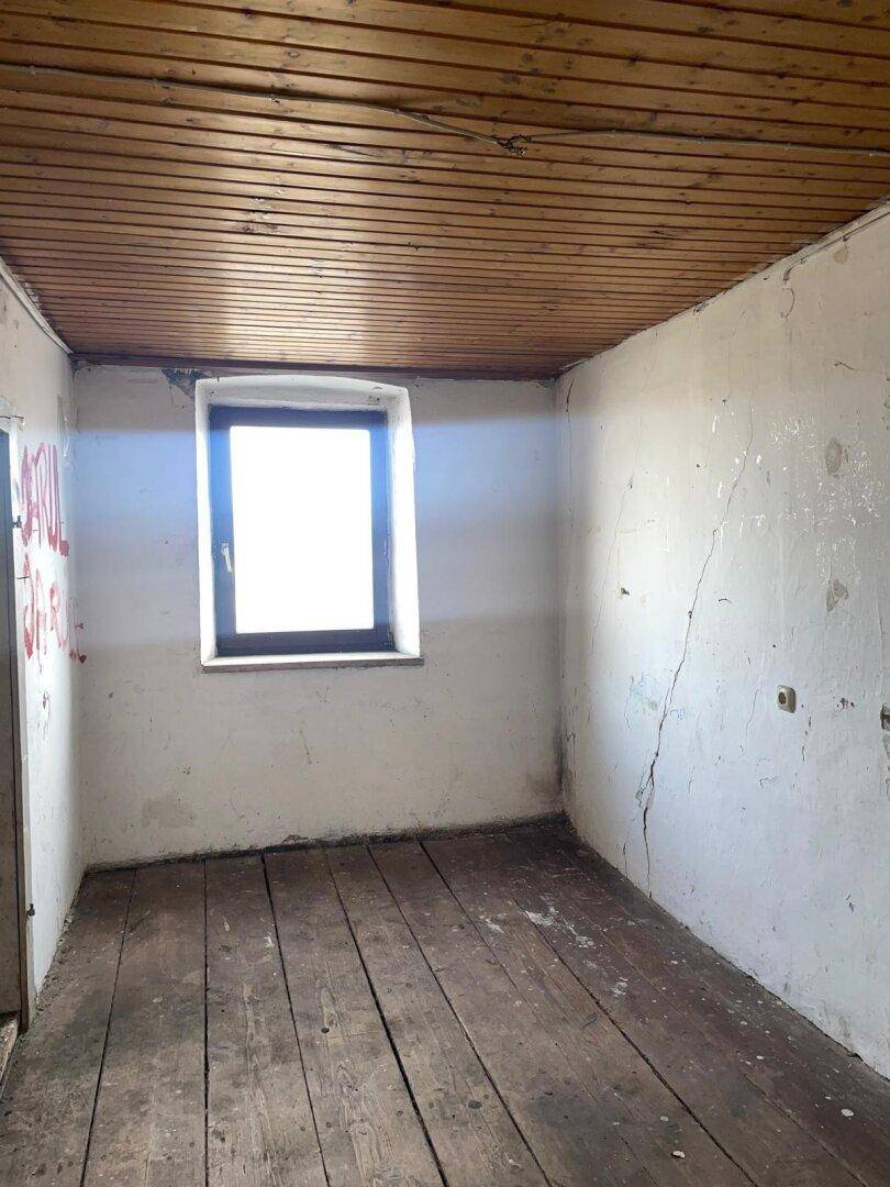 Leerer Raum mit weiß gestrichenen Wänden, Holzdielenboden und einem kleinen Fenster.