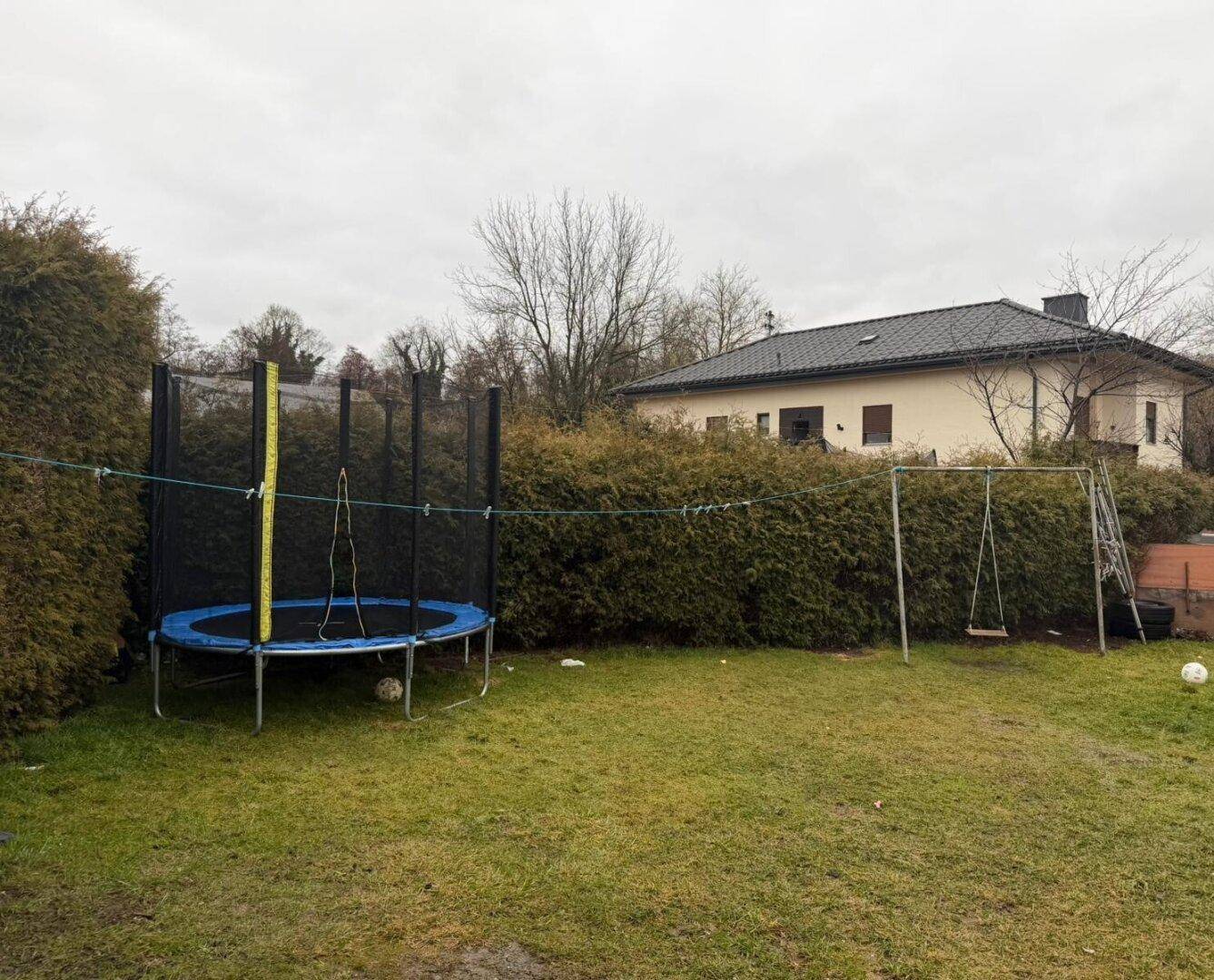 Großer Gartenbereich mit Trampolin und Schaukel für Kinder, umgeben von Hecken.