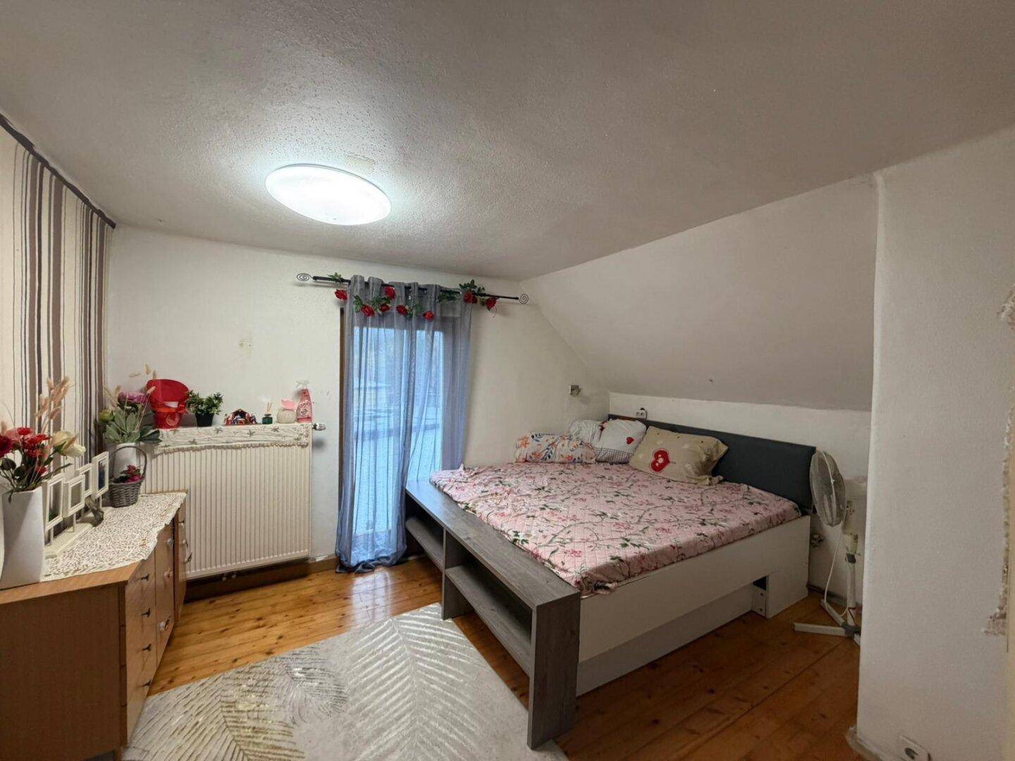 Helles Schlafzimmer im Dachgeschoss mit Holzboden und Fenster, dekoriert mit Blumen.