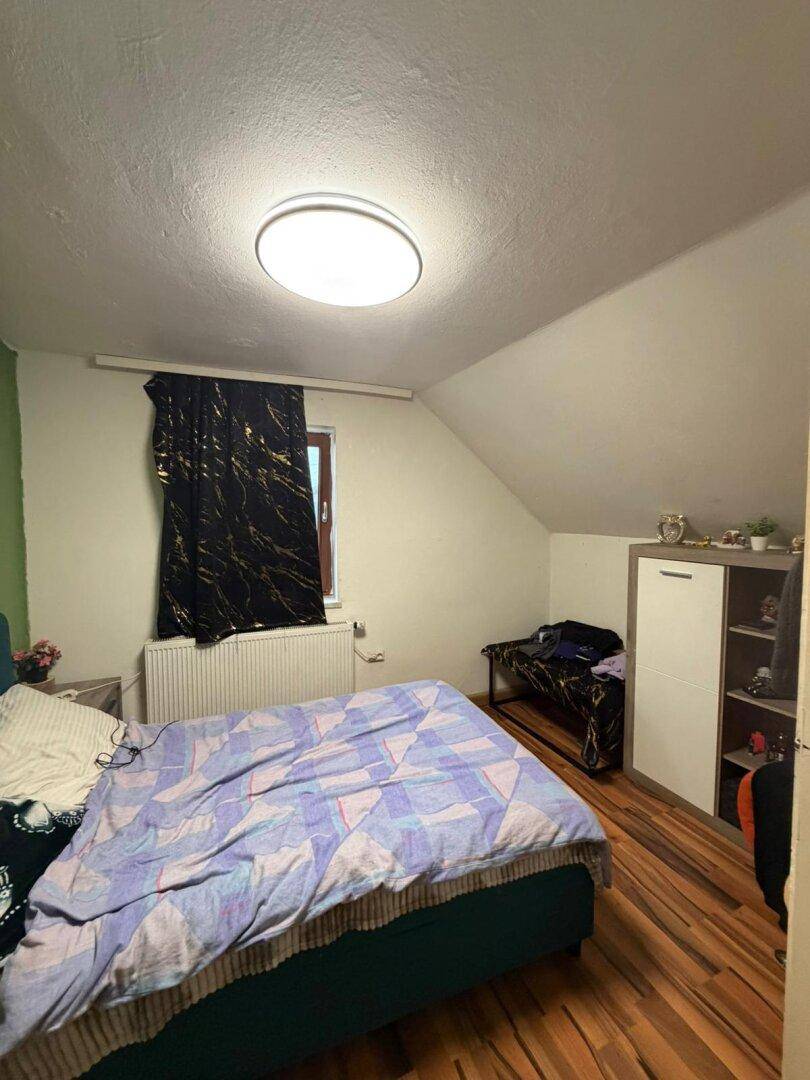 Schlafzimmer im Dachgeschoss mit Holzboden, Heizkörper und dunklen Vorhängen.