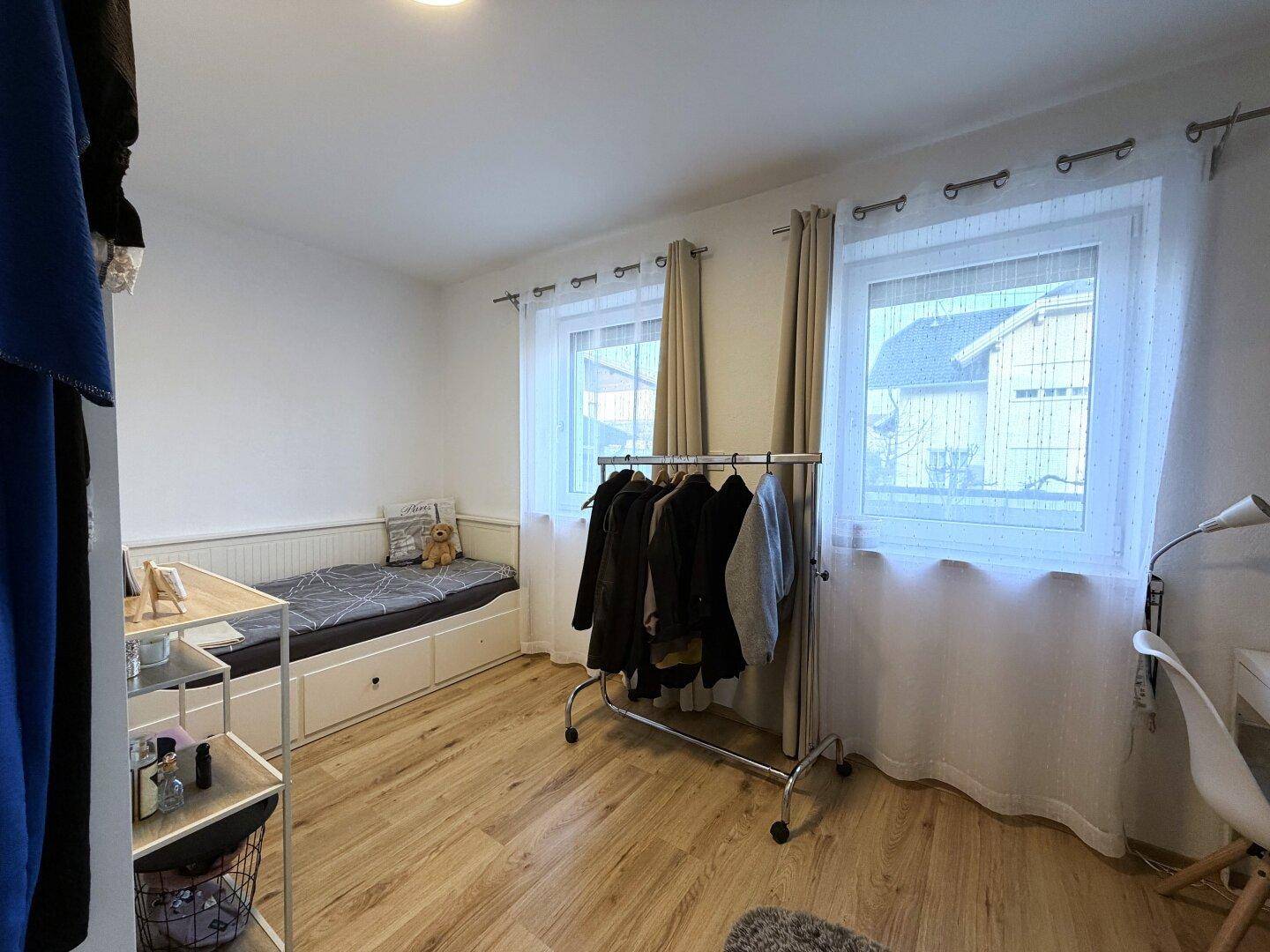 Helles Schlafzimmer mit Holzboden, Tagesbett und Kleiderstange am Fenster.