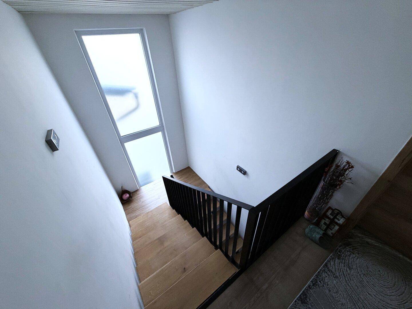 Treppenhaus mit Holzstufen, schwarzem Geländer und hohem Fenster für Tageslicht.
