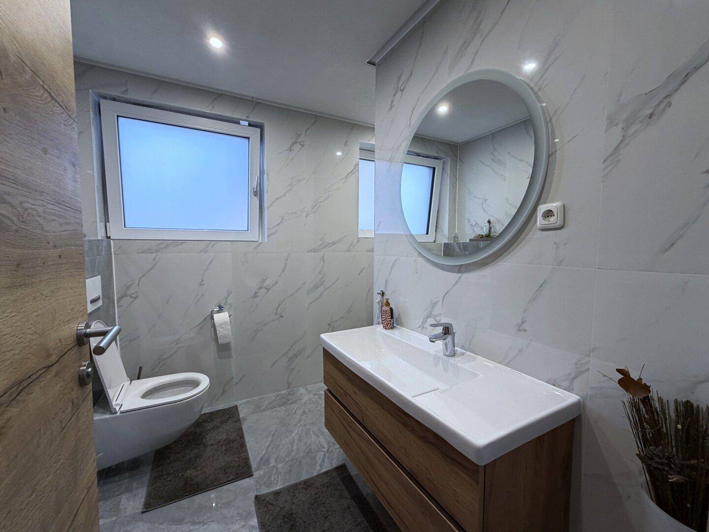 Modernes Badezimmer mit Marmorfliesen, Waschtisch und runden beleuchteten Spiegel.