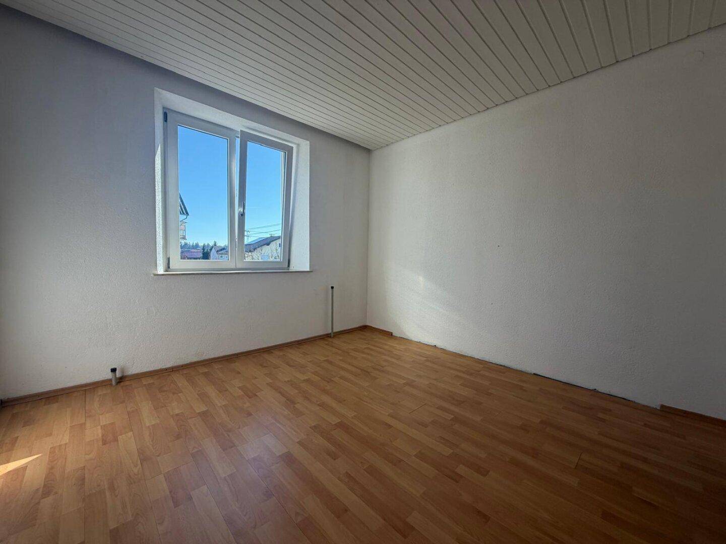 Leeres Schlafzimmer mit Laminatboden und großem Fenster, das viel Tageslicht hereinlässt.