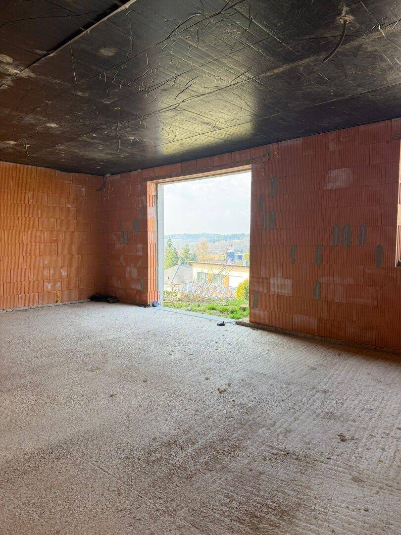 Unfertiger Raum im Rohbau mit Ziegelwänden, Betonboden und großer Fensteröffnung mit Blick ins Grüne.
