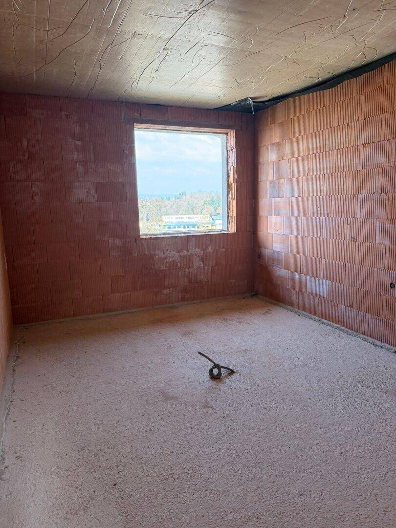Rohbau eines Zimmers mit unverputzten Ziegelwänden, Betonboden und einem Fenster mit Ausblick.