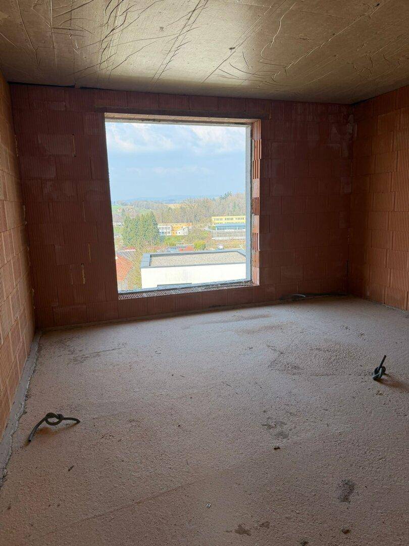 Rohbau eines Zimmers mit unverputzten Ziegelwänden, Betonboden und einem Fenster mit Fernblick.