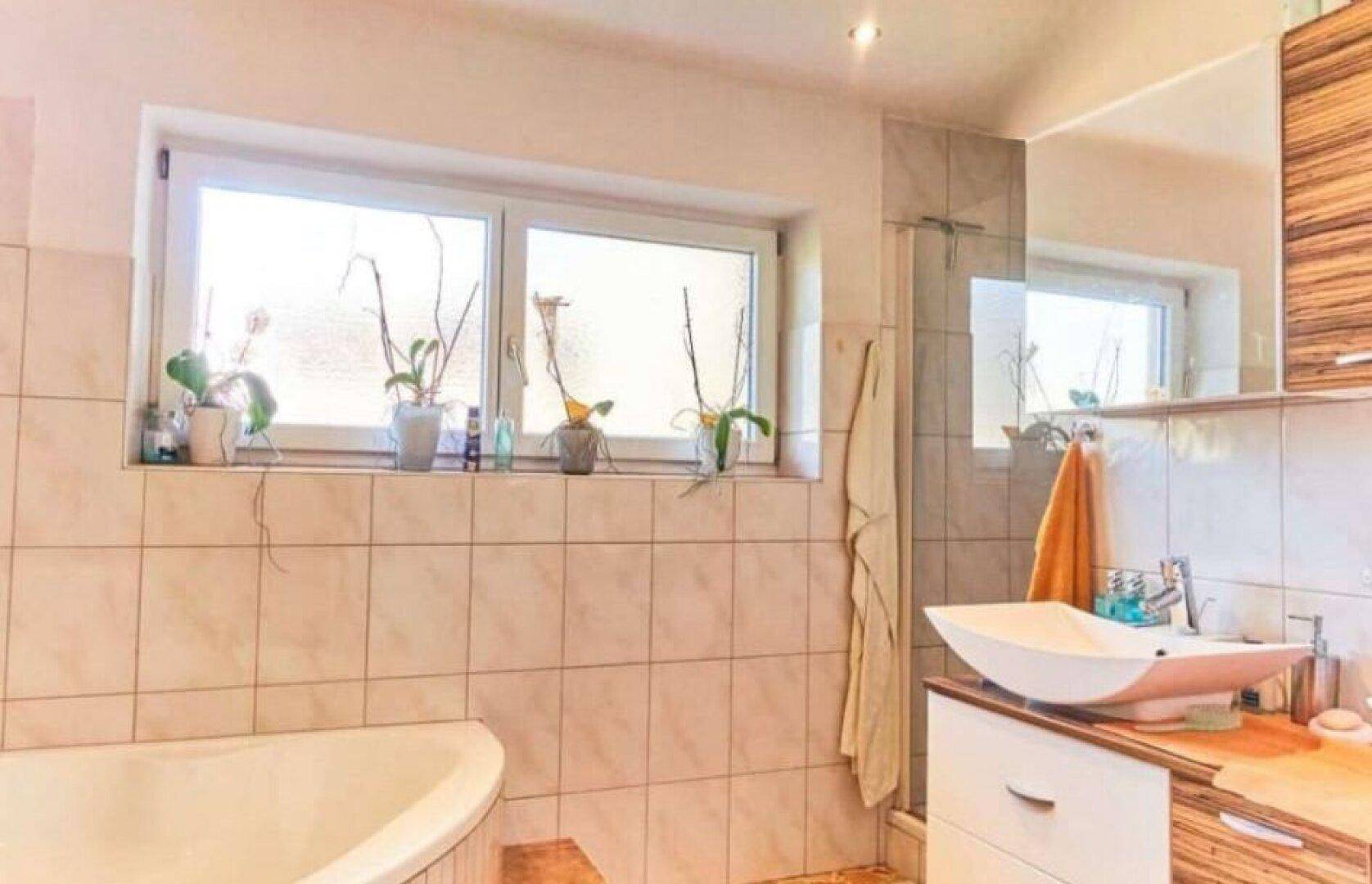 Helles Badezimmer mit Badewanne, Dusche und Waschbecken unter einem großen Fenster.
