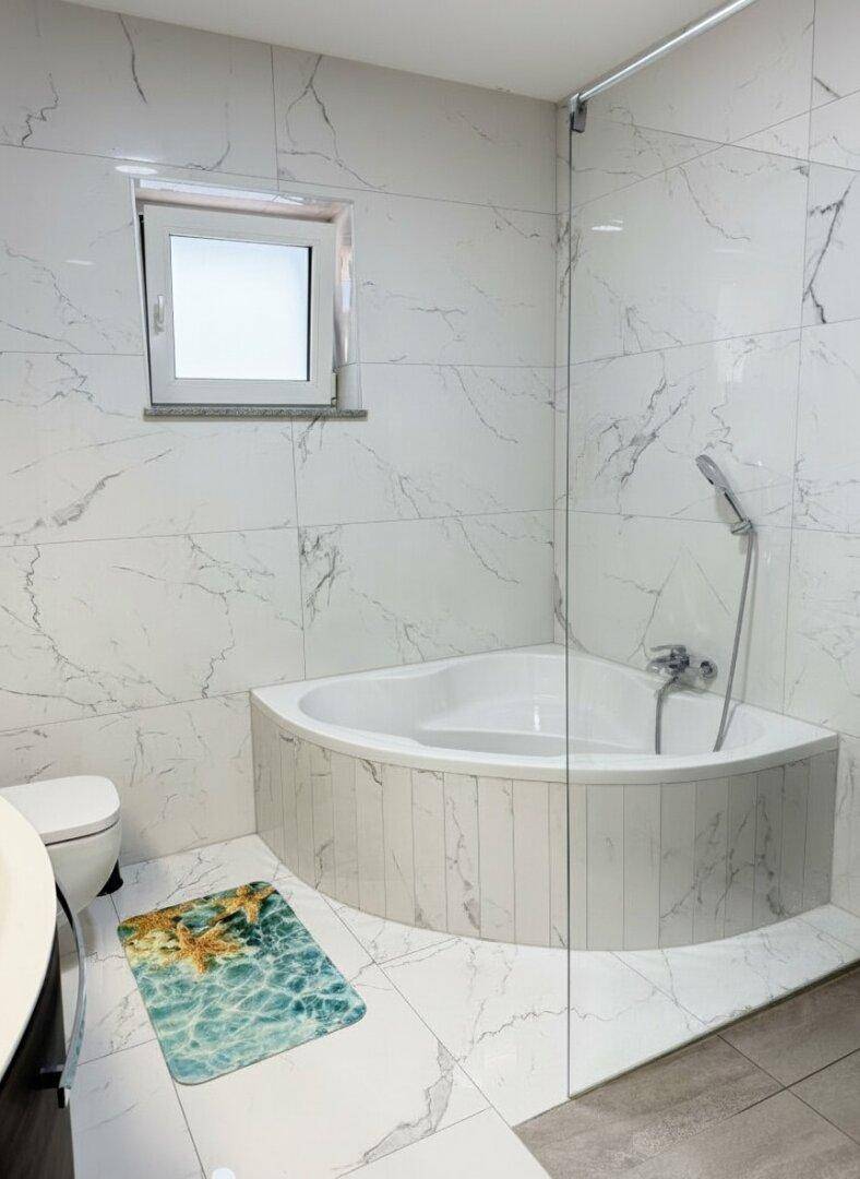 Modernes Badezimmer mit Eckbadewanne, Glasabtrennung und Fenster, komplett gefliest in Marmoroptik.