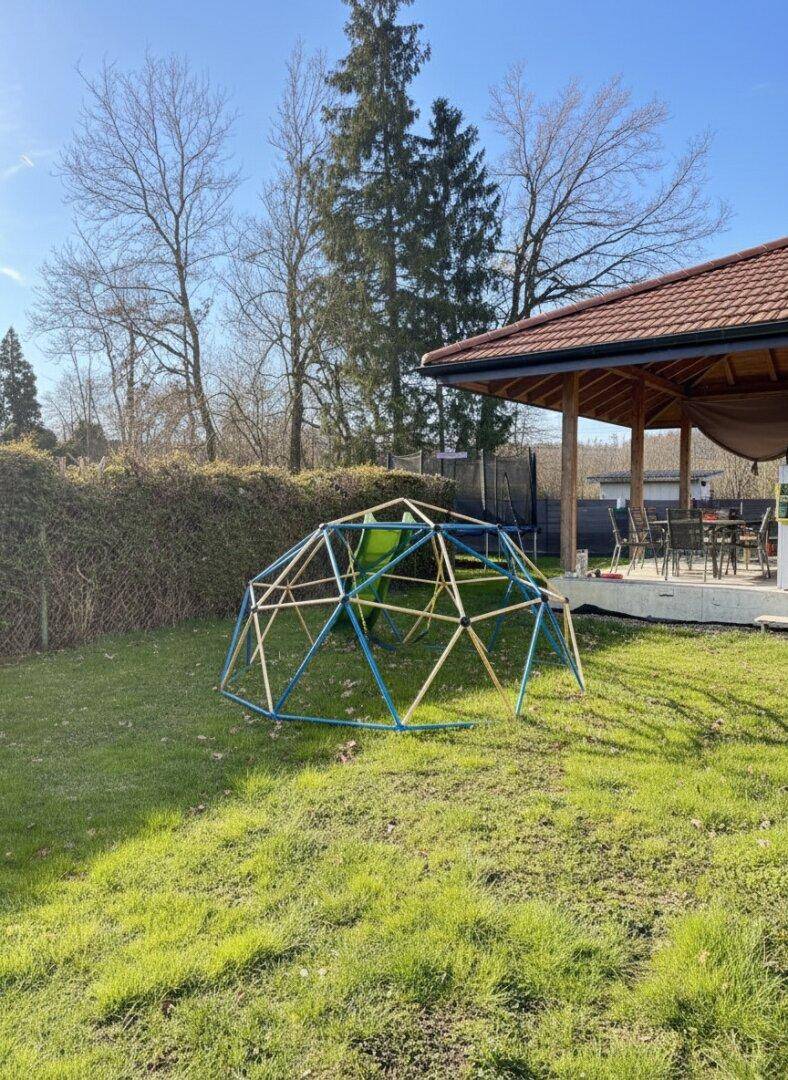 Sonniger Garten mit Klettergerüst auf Rasenfläche und überdachter Terrasse im Hintergrund.