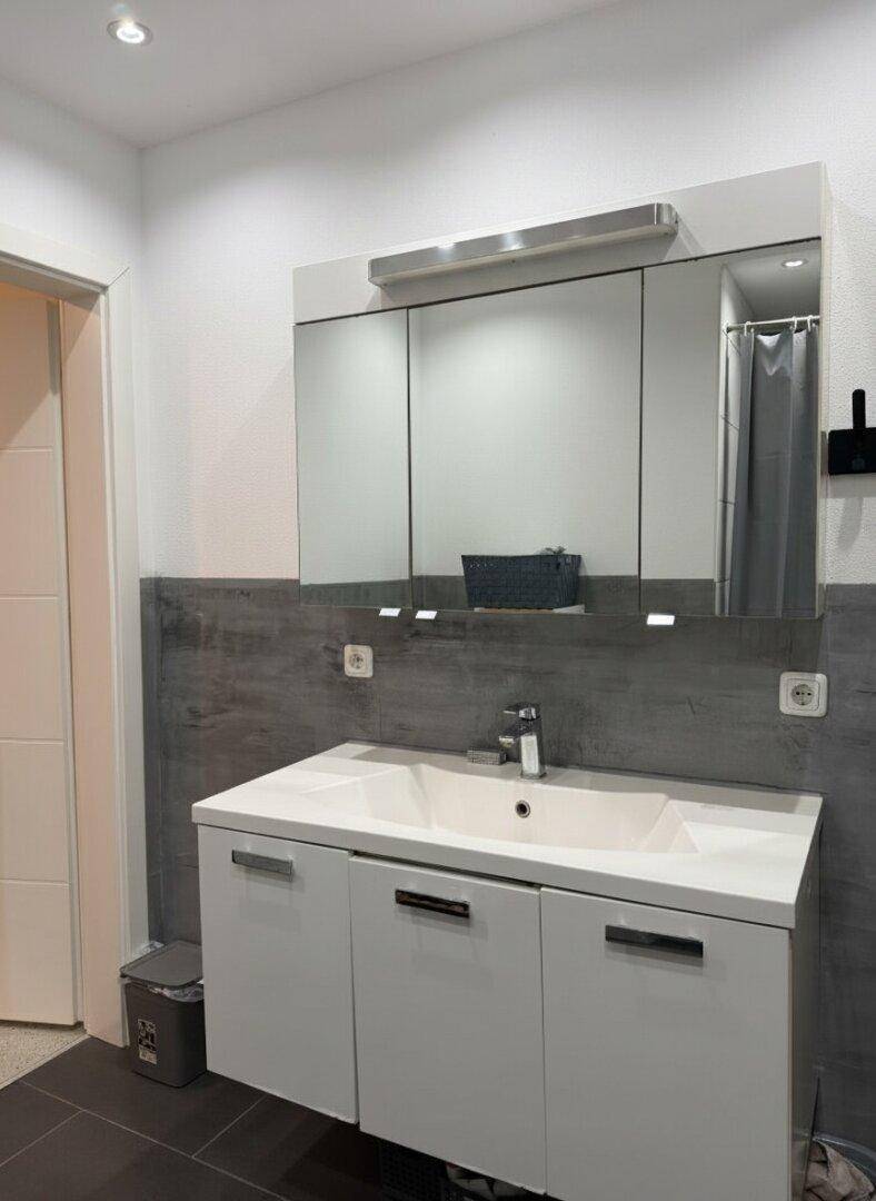 Modernes Badezimmer mit Einzelwaschbecken, Spiegelschrank und dunklen Bodenfliesen.