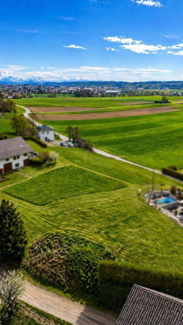 Panoramablick über weite grüne Felder, Häuser und eine Bergkette am Horizont.