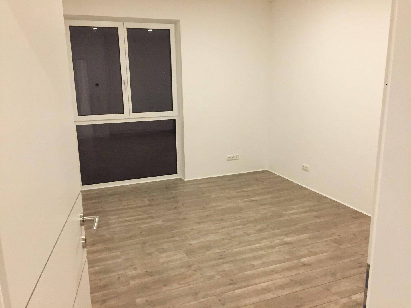 Heller, leerer Raum mit Laminatboden und großem Fenster, bietet viel Tageslicht.