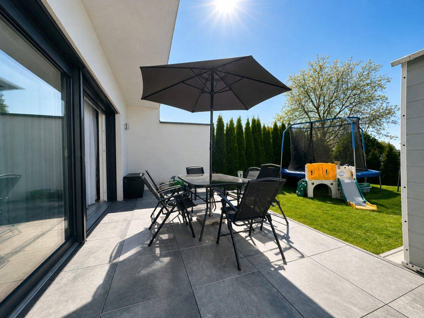 Sonniger Terrassenbereich mit Esstisch, Sonnenschirm und Blick auf den Garten mit Trampolin.
