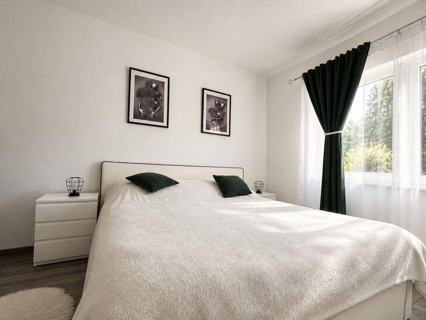 Helles Schlafzimmer mit Doppelbett, Nachttischen und dunklen Vorhängen an einem großen Fenster.