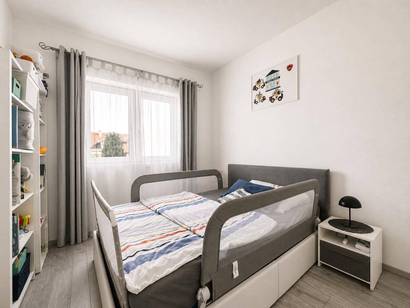 Kinderzimmer mit Einzelbett, Bücherregal und großem Fenster, das viel Tageslicht hereinlässt.