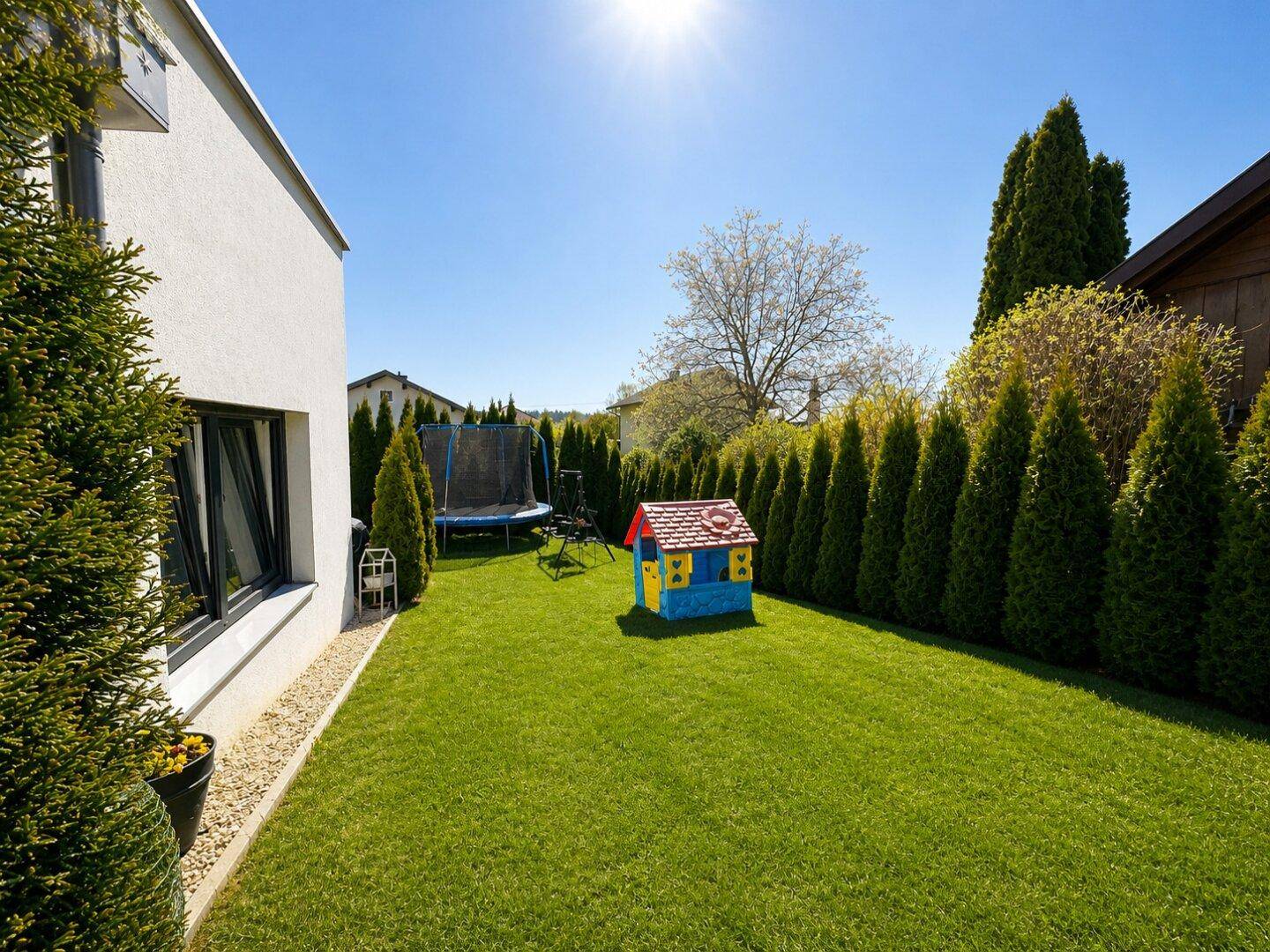 Weitläufiger Garten mit Rasen, Kinderspielhaus und Trampolin, umgeben von hohen Hecken.