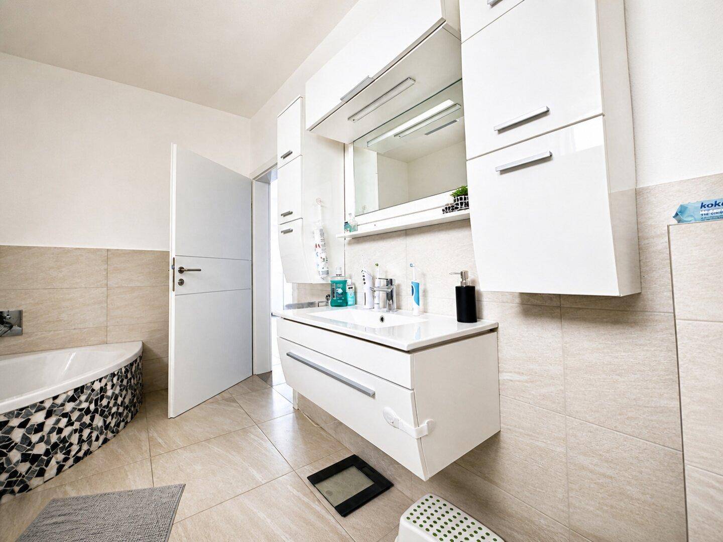 Helles Badezimmer mit modernem Waschtisch, Spiegelschrank und einer Badewanne mit Mosaikfliesen.